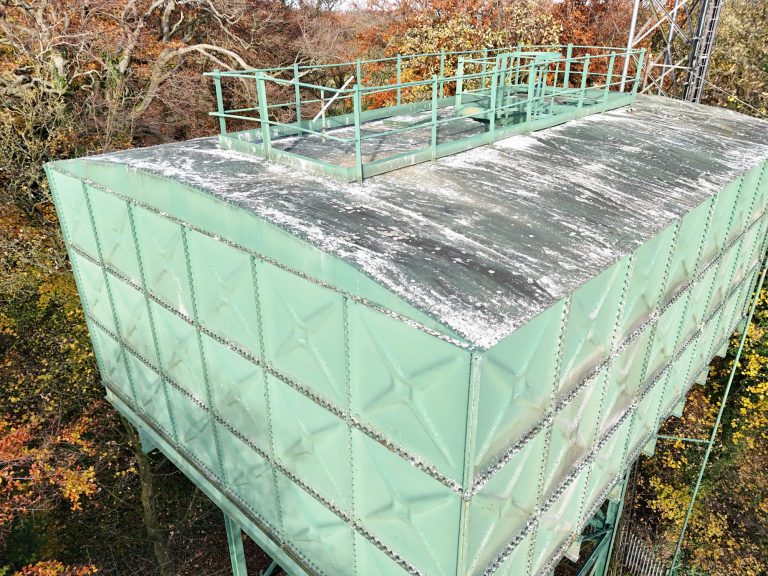 Green industrial water tank with a curved roof, surrounded by trees in autumn foliage.