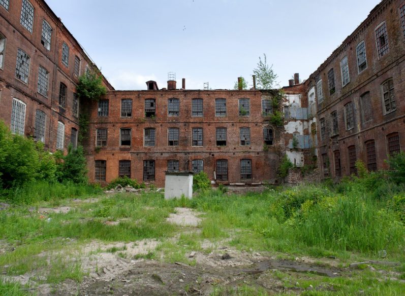 Abandoned red-brick building with broken windows, surrounded by overgrown grass.