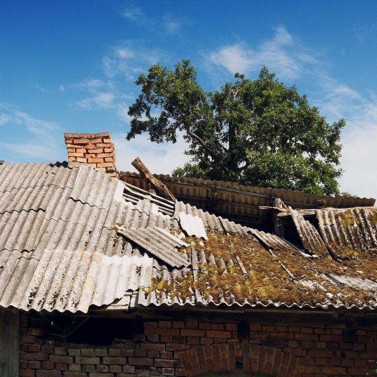 An old building with a damaged, moss-covered asbestos roof under a blue sky.
