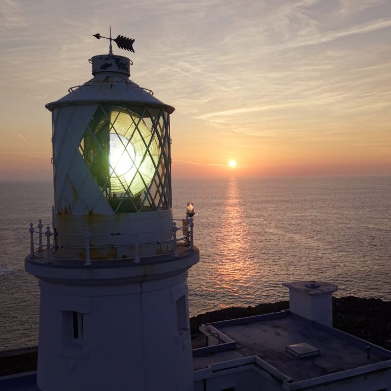 Lighthouse at sunset, with light beam shining across the water and the sun on the horizon.