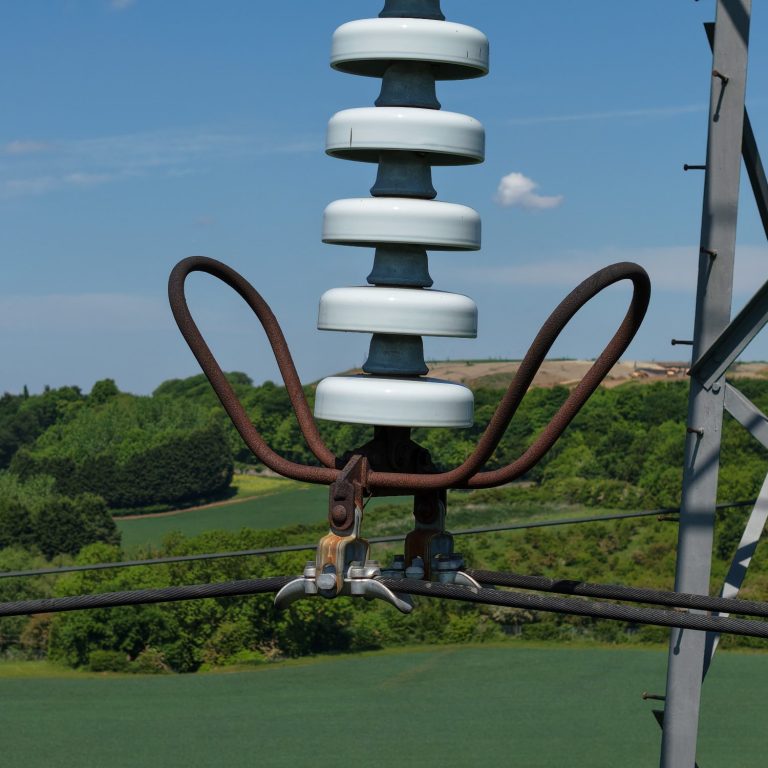 Close-up of an electrical insulator mounted on a metal tower, with green fields in the background.