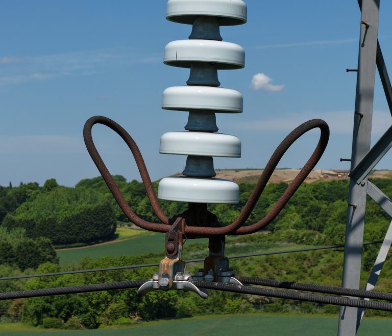 Close-up of an electrical insulator mounted on a metal tower, with green fields in the background.