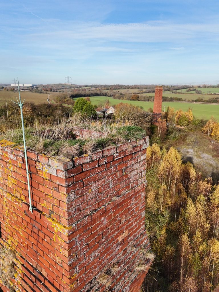 Brick chimney with greenery on top, overlooking a rural landscape with fields and trees.