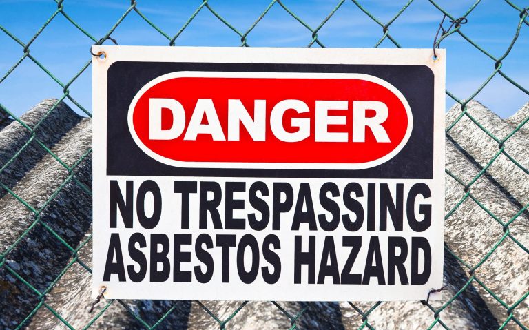 Warning sign stating "Danger: No Trespassing Asbestos Hazard" on a chain-link fence.