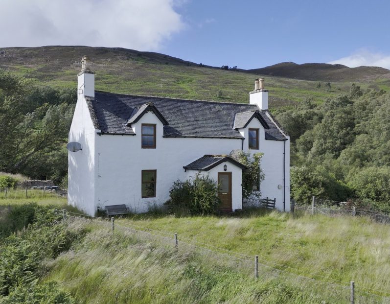 A white cottage surrounded by greenery and hills under a blue sky.