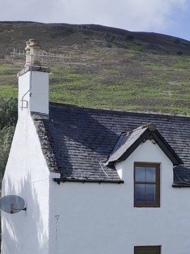 White house with a slate roof, situated at the base of a green, sloping hill.